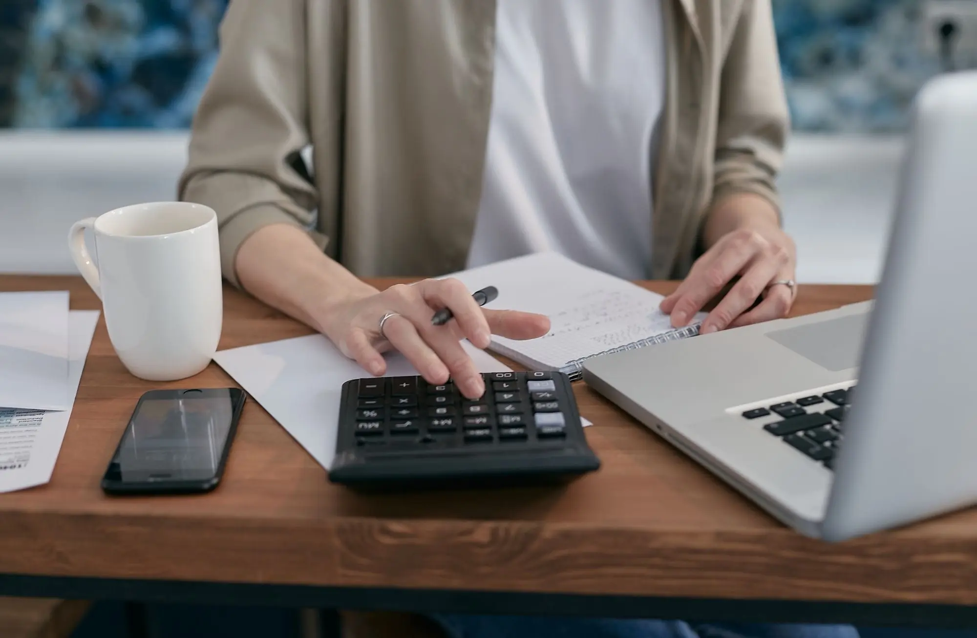 Manos usando una calculadora junto a una laptop, una libreta y una taza